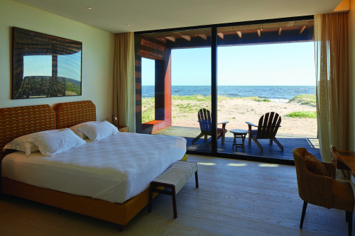 Gurvich Suite iCorten Bungalow Bahía VIK José Ignacio Uruguay bedroom double bed headboard pillows night table lamps chair armchair sand bush sea curtains bench desk sun lights shadow photograph
